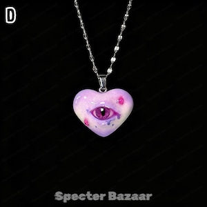 May include: A silver-toned chain necklace featuring a heart-shaped pendant in a light purple hue. The pendant displays a realistic purple eye at its center, accented with small pink and blue spots, and the letter D is visible in the top left corner.