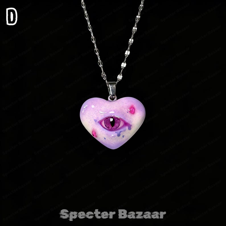 May include: A silver-toned chain necklace featuring a heart-shaped pendant in a light purple hue. The pendant displays a realistic purple eye at its center, accented with small pink and blue spots, and the letter D is visible in the top left corner.