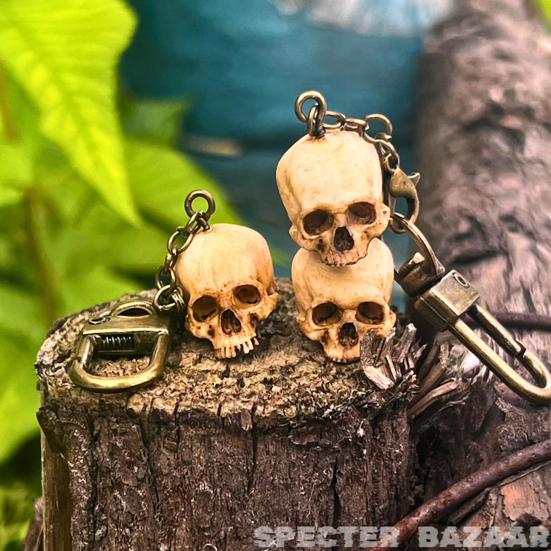 May include: Three small, detailed, bone-colored skull keychains with bronze rings and chains. The skulls are stacked on top of each other, with the top skull facing forward and the bottom two facing slightly to the side. The keychains are on a wooden surface.