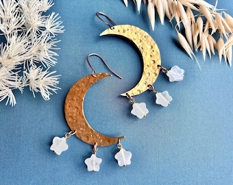 May include: A pair of brass crescent moon earrings with a hammered texture. Each earring has three dangling star-shaped, white gemstone charms. The earrings are set against a blue background with dried floral accents.