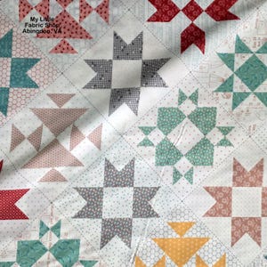 Puede incluir: Primer plano de un edredón de patchwork con varios patrones geométricos. El edredón presenta diseños de estrellas y triángulos en tonos de rojo, verde azulado, gris y amarillo, sobre un fondo blanco. El texto "My Little Fabric Shop Abingdon, VA" es visible.