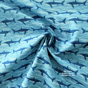 May include: Light blue fabric with a repeating pattern of dark blue sharks swimming in wavy lines. The fabric is folded, showing the texture and design. The overall theme is marine life.