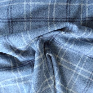 Puede incluir: Primer plano de una tela azul claro con un patrón de cuadros. La tela tiene un aspecto suave y texturizado con una cuadrícula de líneas blancas y azul oscuro. La tela está doblada, creando sombras y profundidad.