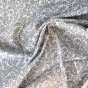 May include: White fabric with a repeating pattern of small, light blue leafy branches. The fabric is folded in a way that shows the pattern repeating.