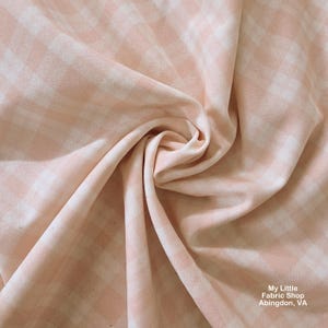 May include: A close-up shot of a pink and white gingham fabric, showing the woven pattern. The fabric is folded, revealing its texture and drape. This material is likely suitable for dressmaking or crafting projects.