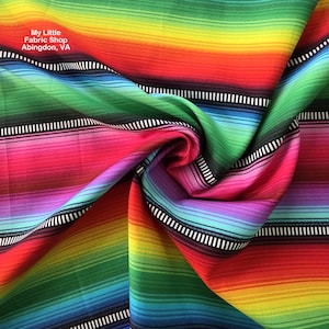 Op de afbeelding: Een levendige stof met een regenboogstreepmotief. De stof heeft horizontale banden in rood, oranje, geel, groen, blauw en paars. Zwart-witte streepaccenten zijn ook aanwezig. De tekst "My Little Fabric Shop Abingdon, VA" is zichtbaar.