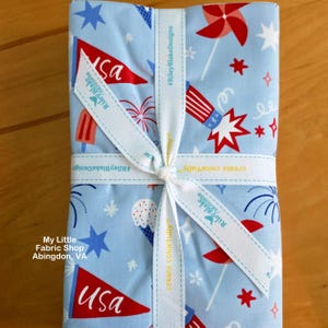 May include: A bundle of light blue fabric with a patriotic print, featuring red, white, and blue stars, fireworks, and flags. The fabric is tied with a white ribbon that reads "Riley Blake Designs" and "create colorfully."