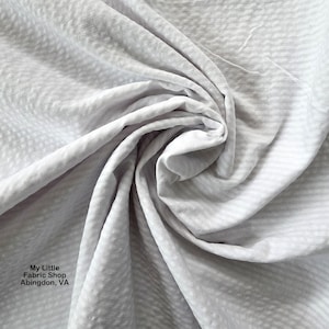 Puede incluir: Primer plano de una tela gris claro con un patrón texturizado y ligeramente elevado. La tela está arrugada y doblada, creando sombras y reflejos. El texto "My Little Fabric Shop Abingdon, VA" es visible en la esquina inferior izquierda.