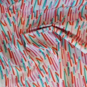 以下が含まれることがあります： 白地に、ピンク、オレンジ、ターコイズの色のブラシストロークのようなカラフルな線が繰り返しパターンで描かれた生地。生地には、薄い白いドット模様があります。