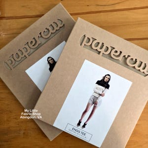 May include: Two square tan cardboard envelopes featuring the die-cut word papercut at the top. Each envelope displays a white rectangular label with a photograph of a person wearing a black and white Ensis Tee and gray shorts, with the text Ensis Tee sizes XXS XS S M L XL.