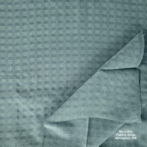 Puede incluir: Primer plano de una tela color verde azulado con un patrón texturizado en forma de cuadrícula. Una esquina de la tela está doblada, revelando el reverso. La tela parece ser de peso medio, adecuada para varios proyectos de costura. El texto "My Little Fabric Shop Abingdon, VA" es visible.