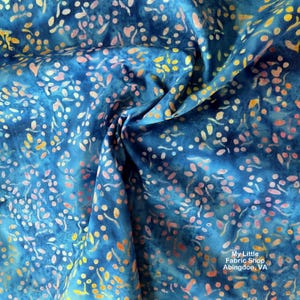 Può includere: Primo piano di un tessuto blu con un design batik. Il tessuto presenta un motivo di piccole forme irregolari in tonalità di rosa, giallo e bianco, sparse sullo sfondo blu. Il tessuto è leggermente stropicciato, creando ombre e profondità.