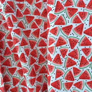 May include: White fabric featuring a repeating pattern of watermelon slices. The watermelons are red with black seeds and green rinds. The fabric is likely cotton or a similar material, suitable for crafting or clothing.