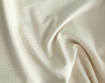 May include: A close-up of a cream-colored fabric with a subtle cross-stitch pattern. The fabric is soft and textured, with a slightly wrinkled appearance.