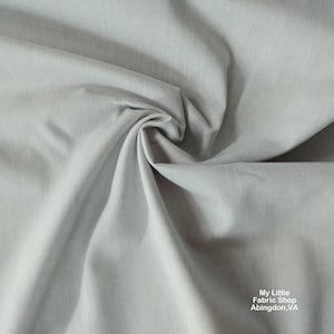 May include: A close-up of a light gray fabric with a smooth texture. The fabric is folded and draped, showing its soft and flowing nature.
