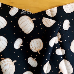 Puede incluir: Tela negra con un patrón repetido de calabazas blancas con detalles dorados y pequeños puntos dorados. La tela está doblada, mostrando el patrón desde diferentes ángulos. El texto "My Little Fabric Shop Abingdon, Va" es visible.