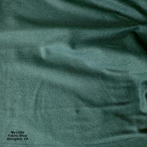 May include: Close-up of a dark green fabric with a soft, textured surface. The fabric has subtle folds and shadows, creating depth. The image includes the text "My Little Fabric Shop Abingdon, VA".