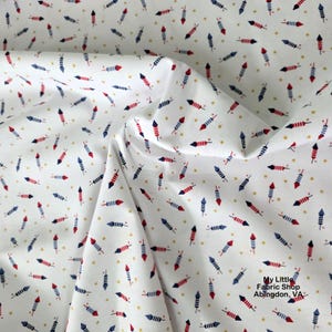 Könnte beinhalten: Weißer Stoff mit einem sich wiederholenden Muster aus roten, blauen und weißen Feuerwerkskörpern und kleinen goldenen Sternen. Der Stoff ist gefaltet und zeigt die Textur und den Fall des Materials. Der Text "My Little Fabric Shop Abingdon, VA" ist sichtbar.