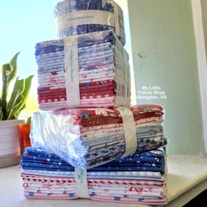 Może przedstawiać: Stos pakietów tkanin owiniętych w przezroczysty plastik z białymi papierowymi opaskami z napisem Riley Blake Designs. Tkanina posiada czerwone, białe i niebieskie wzory, w tym gwiazdki, paski i motywy kwiatowe. Tekst po prawej stronie brzmi My Little Fabric Shop Abingdon, VA.