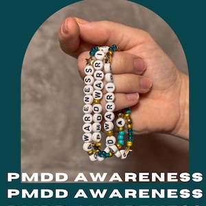May include: Three beaded bracelets featuring white letter beads spelling PMDD AWARENESS and WARRIOR, interspersed with metallic gold spherical beads, small teal blue translucent beads, and gold-toned star-shaped charms on a flexible elastic cord.