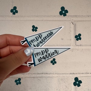 May include: Two triangular pennant stickers with a light blue background and black borders. The top sticker features the black text PMDD Awareness, and the bottom sticker features the black text PMDD warrior. Both stickers have small black tassels.