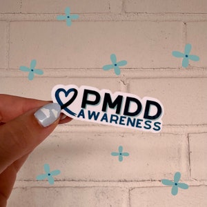 May include: A white die-cut sticker featuring a dark blue awareness ribbon shaped into a heart on the left side. The text PMDD is printed in large, bold, dark blue letters, with the word AWARENESS in smaller dark blue letters underneath.