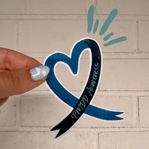 May include: A die-cut sticker shaped like a heart formed by a ribbon in shades of dark blue and teal. The text PMDD Awareness is printed in a teal script font along the right side of the ribbon. Above the heart are three teal brush strokes.