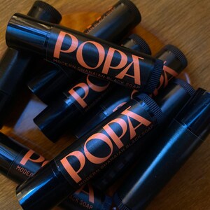 Puede incluir: Primer plano de varios tubos de bálsamo labial negros con texto naranja que dice "POPA MODERN GHANAIAN BLACK SOAP".