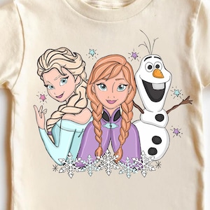 Puede incluir: Una camiseta color crema con un gr&aacute;fico de Elsa, Anna y Olaf de la pel&iacute;cula Frozen. Elsa lleva un vestido azul claro, Anna un vestido morado y Olaf es un mu&ntilde;eco de nieve. El dise&ntilde;o incluye copos de nieve y estrellas.