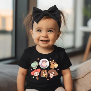 May include: A smiling baby wearing a black t-shirt featuring cartoon character faces from the movie Toy Story. The shirt has a round neck and short sleeves. The baby is also wearing a black bow headband.