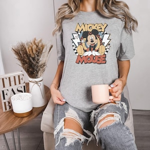May include: A gray t-shirt featuring a vintage-style graphic of Mickey Mouse. The design includes the words "Mickey Mouse" in a retro font, with Mickey sitting in a relaxed pose. The shirt is paired with ripped jeans.
