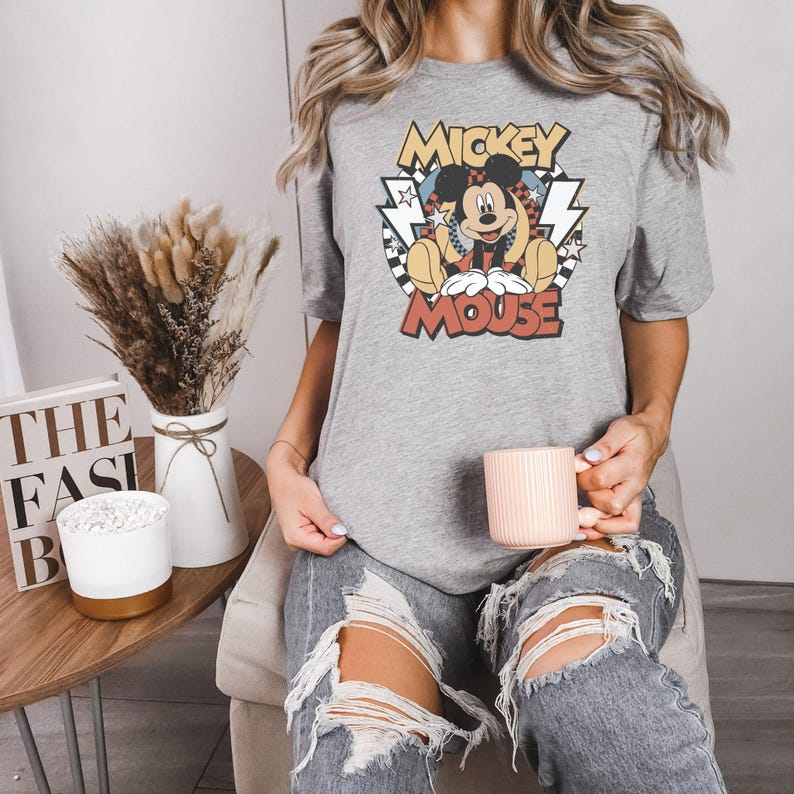 May include: A gray t-shirt featuring a vintage-style graphic of Mickey Mouse. The design includes the words "Mickey Mouse" in a retro font, with Mickey sitting in a relaxed pose. The shirt is paired with ripped jeans.