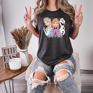 Puede incluir: Camiseta gris oscuro con Elsa, Anna y Olaf de Frozen. El gr&aacute;fico incluye los personajes con copos de nieve y estrellas. La camiseta la lleva una persona con vaqueros azules rotos.
