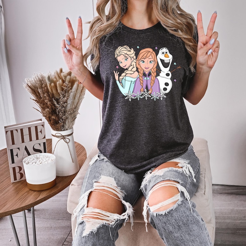 Puede incluir: Camiseta gris oscuro con Elsa, Anna y Olaf de Frozen. El gr&aacute;fico incluye los personajes con copos de nieve y estrellas. La camiseta la lleva una persona con vaqueros azules rotos.
