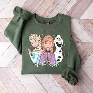 Puede incluir: Una sudadera verde oliva con un gr&aacute;fico de los personajes animados Elsa, Anna y Olaf. Elsa lleva un vestido azul claro, Anna un vestido morado y Olaf es un mu&ntilde;eco de nieve. El dise&ntilde;o incluye copos de nieve y estrellas.