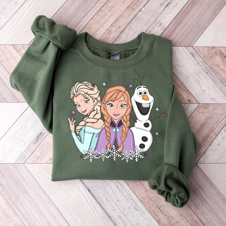 Puede incluir: Una sudadera verde oliva con un gr&aacute;fico de los personajes animados Elsa, Anna y Olaf. Elsa lleva un vestido azul claro, Anna un vestido morado y Olaf es un mu&ntilde;eco de nieve. El dise&ntilde;o incluye copos de nieve y estrellas.