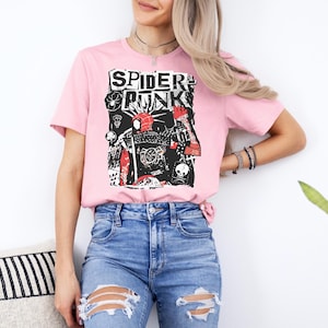 May include: A light pink t-shirt featuring a graphic design with the words "SPIDER PUNK" in a bold, black font. The graphic includes a stylized image of a spider and other punk-inspired elements. The shirt is paired with blue jeans.