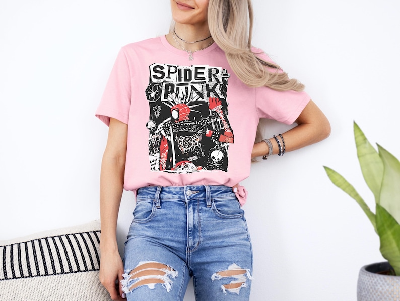 May include: A light pink t-shirt featuring a graphic design with the words "SPIDER PUNK" in a bold, black font. The graphic includes a stylized image of a spider and other punk-inspired elements. The shirt is paired with blue jeans.