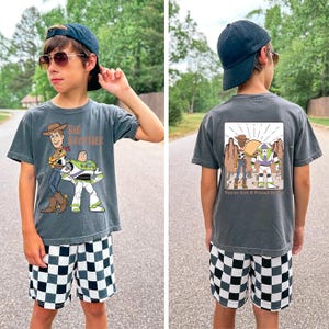 Op de afbeelding: Een antracietgrijs t-shirt met een cartoonafbeelding van Woody en Buzz Lightyear, met de tekst "Big Brother". De achterkant van de shirt toont dezelfde personages met de tekst "You've Got A Friend In Me". De shorts zijn zwart-wit geruit.