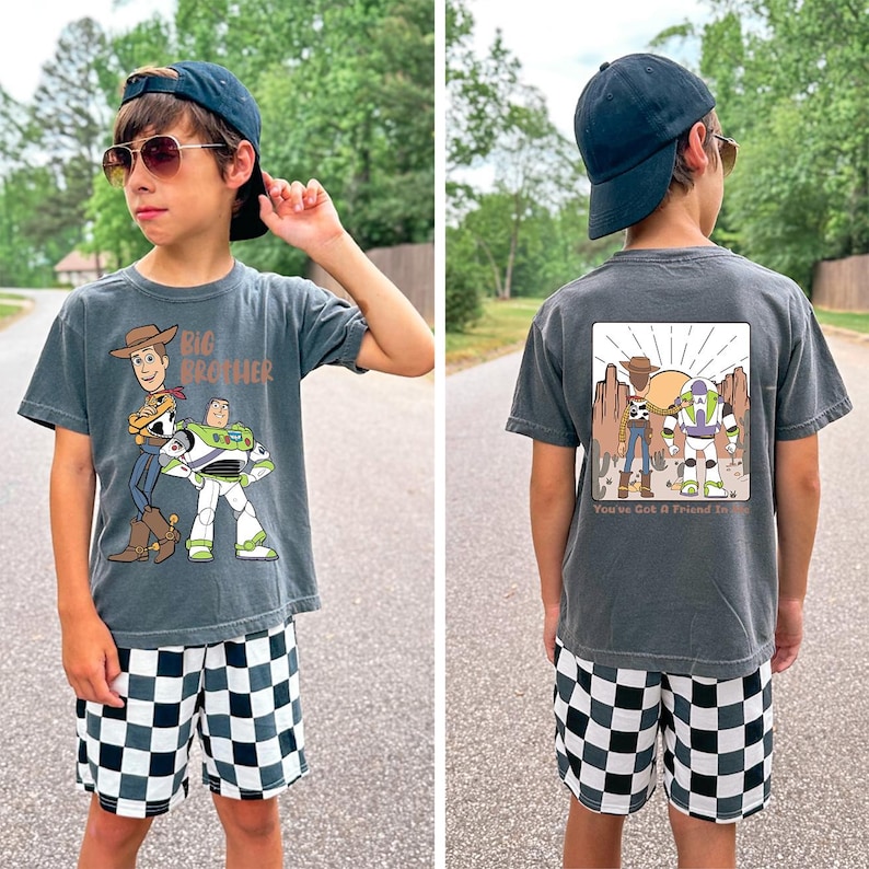 Op de afbeelding: Een antracietgrijs t-shirt met een cartoonafbeelding van Woody en Buzz Lightyear, met de tekst "Big Brother". De achterkant van de shirt toont dezelfde personages met de tekst "You've Got A Friend In Me". De shorts zijn zwart-wit geruit.