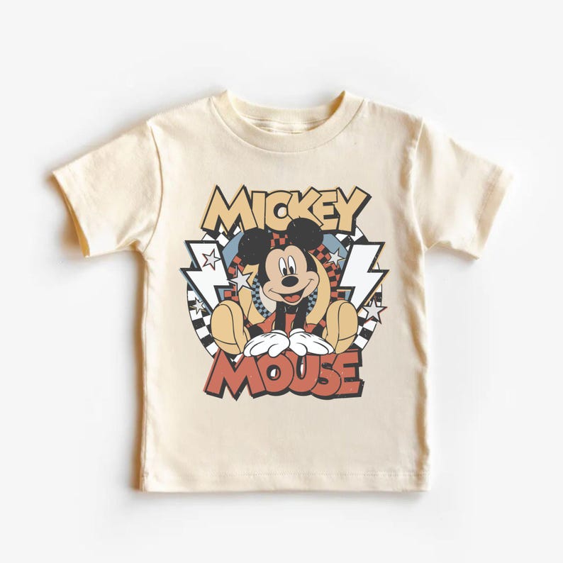 May include: A cream-colored t-shirt featuring a graphic of Mickey Mouse. The design includes the words "Mickey Mouse" in a retro font, with lightning bolts and a checkered background. The shirt is short-sleeved.