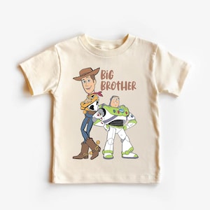 May include: Cream-colored t-shirt featuring cartoon characters Woody and Buzz Lightyear from Toy Story. The words "BIG BROTHER" are printed in brown. Woody is wearing a cowboy hat, and Buzz is in a spacesuit.