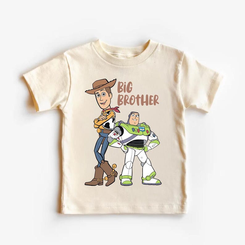 Op de afbeelding: Cr&egrave;mekleurig T-shirt met de tekenfilmfiguren Woody en Buzz Lightyear uit Toy Story. De woorden "BIG BROTHER" zijn in bruin gedrukt. Woody draagt een cowboyhoed en Buzz een ruimtepak.