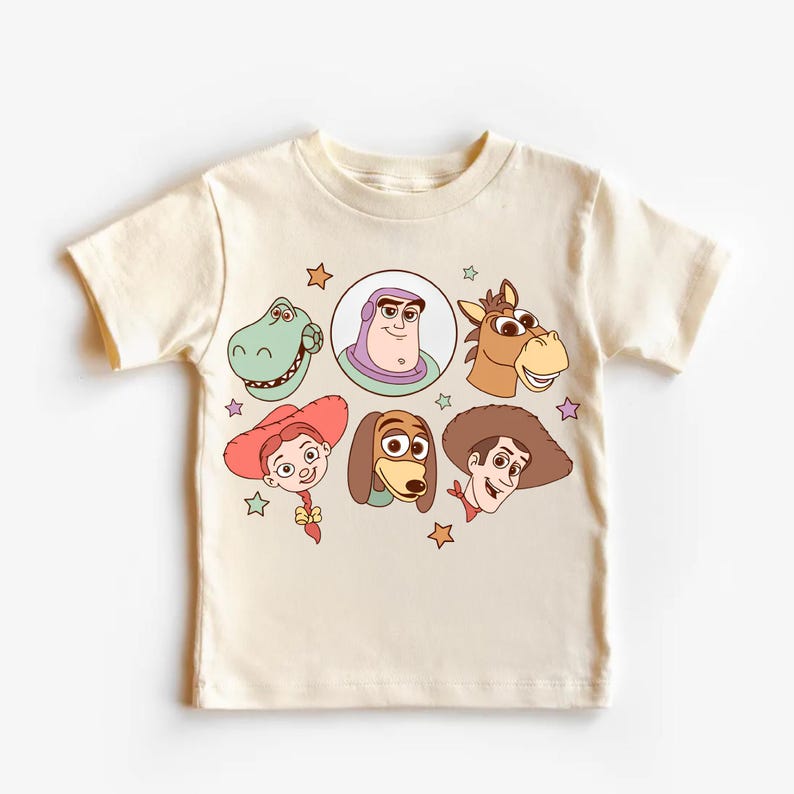 May include: Cream-colored t-shirt featuring cartoon character faces from the Toy Story movie. The design includes a green dinosaur, Buzz Lightyear, a horse, Jessie, Slinky Dog, and Woody, with star accents.
