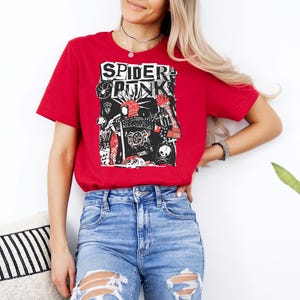 May include: Red t-shirt with the words "SPIDER PUNK" in a bold, distressed font. The graphic features a collage of illustrations in black, white, and red, including a skull and a spider web. The shirt is paired with blue jeans.