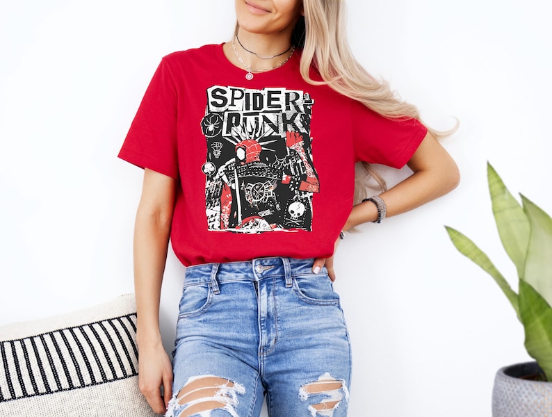 May include: Red t-shirt with the words "SPIDER PUNK" in a bold, distressed font. The graphic features a collage of illustrations in black, white, and red, including a skull and a spider web. The shirt is paired with blue jeans.
