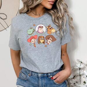 May include: A heather gray t-shirt featuring a colorful graphic of cartoon characters. The design includes images of a green dinosaur, Buzz Lightyear, a horse, a cowgirl, a dog, and a cowboy. The shirt is made of a soft material.