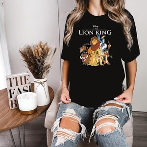May include: Black t-shirt featuring a graphic of characters from Disney's The Lion King, including Simba, Timon, Pumbaa, and Mufasa. The text "Disney The Lion King" is printed above the graphic. The shirt is a crew neck style.