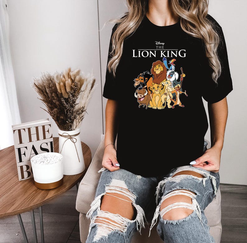 May include: Black t-shirt featuring a graphic of characters from Disney's The Lion King, including Simba, Timon, Pumbaa, and Mufasa. The text "Disney The Lion King" is printed above the graphic. The shirt is a crew neck style.