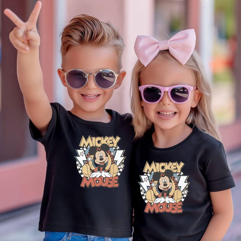 May include: Two children wearing black t-shirts with a Mickey Mouse graphic. The graphic includes the words "Mickey Mouse" and an image of Mickey Mouse. One child wears sunglasses and makes a peace sign. The other child wears a pink bow and sunglasses.